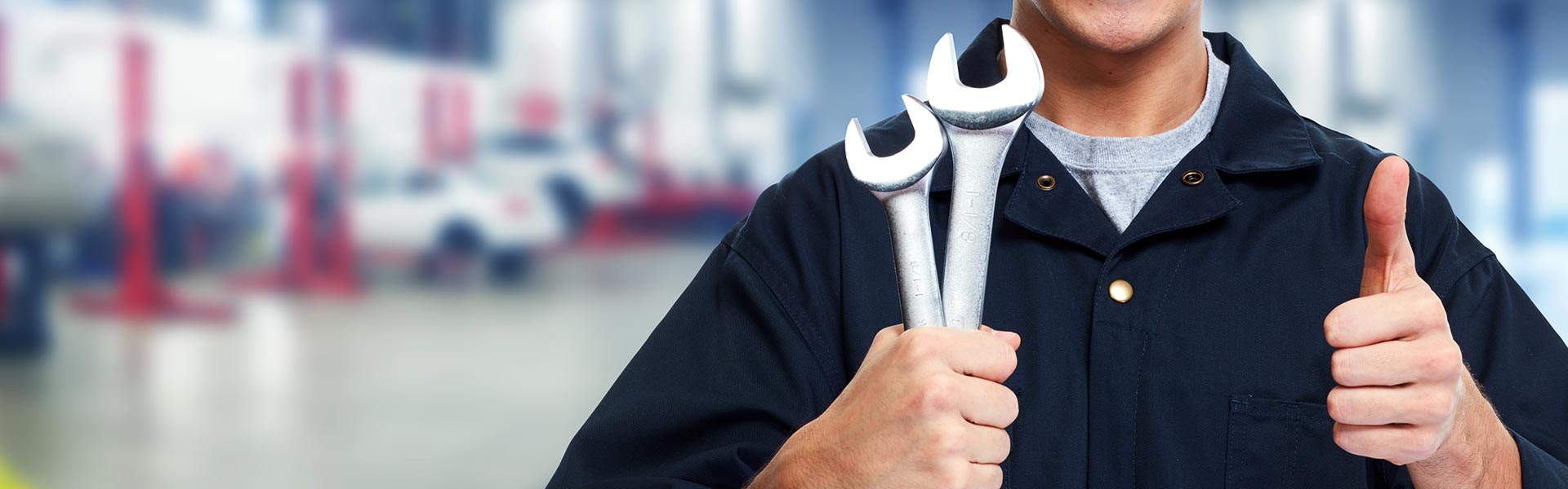 Automotive service technician holding wrenches and giving a thumbs-up inside a modern auto service center.
